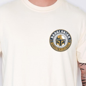 May include: A cream-colored short-sleeve t-shirt featuring a circular graphic on the left chest. The design includes a gold and black helmet icon with the text DADALORIAN, THIS IS THE WAY, and NUMBER ONE IN THE GALAXY printed in a circular border.