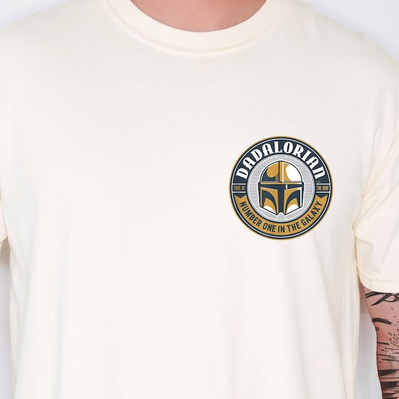 May include: A cream-colored short-sleeve t-shirt featuring a circular graphic on the left chest. The design includes a gold and black helmet icon with the text DADALORIAN, THIS IS THE WAY, and NUMBER ONE IN THE GALAXY printed in a circular border.