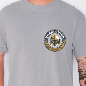 May include: A light gray t-shirt featuring a circular graphic on the left chest. The design includes a gold and black helmet icon, the text DADALORIAN at the top, and the text NUMBER ONE IN THE GALAXY at the bottom, with THIS IS THE WAY on the sides.