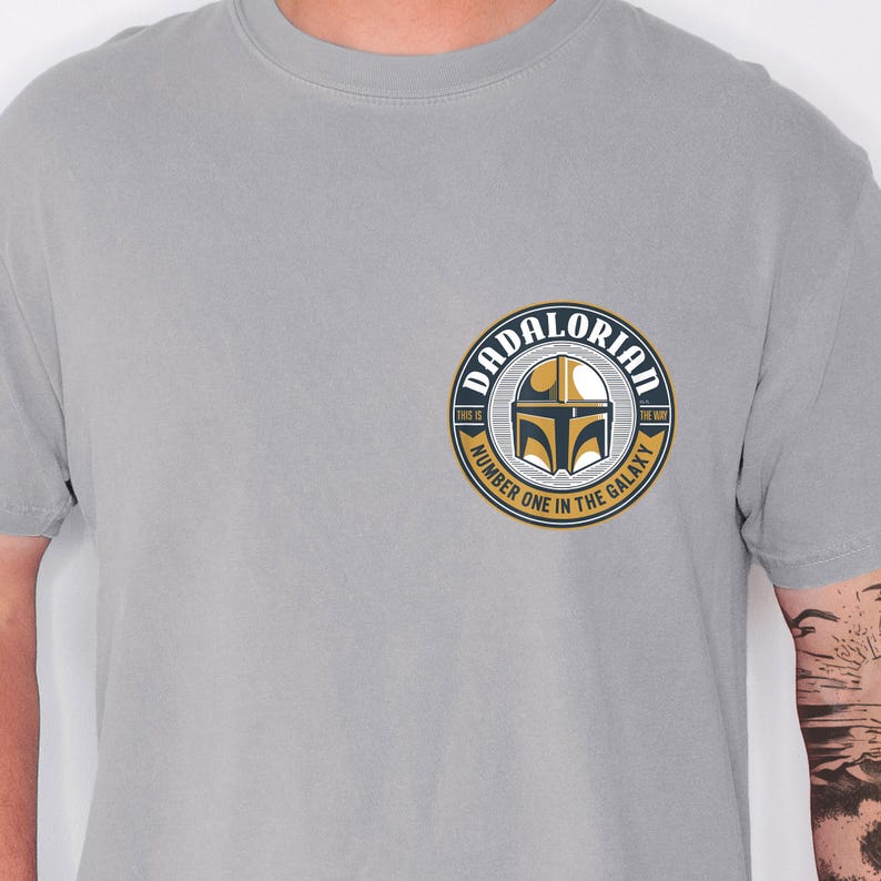 May include: A light gray t-shirt featuring a circular graphic on the left chest. The design includes a gold and black helmet icon, the text DADALORIAN at the top, and the text NUMBER ONE IN THE GALAXY at the bottom, with THIS IS THE WAY on the sides.