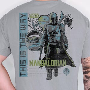 May include: A grey t-shirt featuring a graphic print of The Mandalorian and Grogu. The design includes the text "THIS IS THE WAY" and "THE MANDALORIAN" alongside Star Wars imagery, including TIE fighters and a small circular planet graphic.