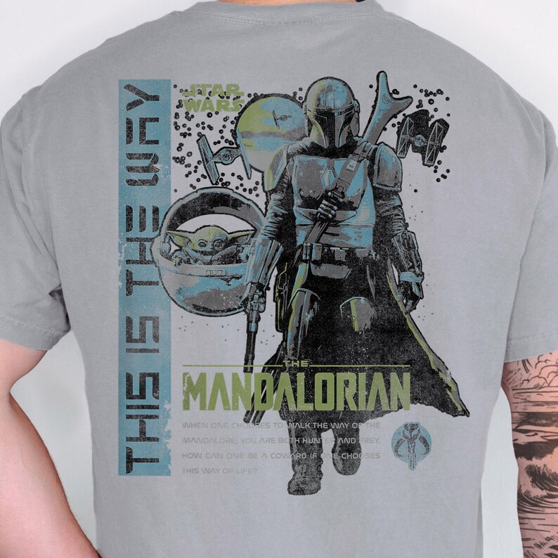 May include: A grey t-shirt featuring a graphic print of The Mandalorian and Grogu. The design includes the text "THIS IS THE WAY" and "THE MANDALORIAN" alongside Star Wars imagery, including TIE fighters and a small circular planet graphic.
