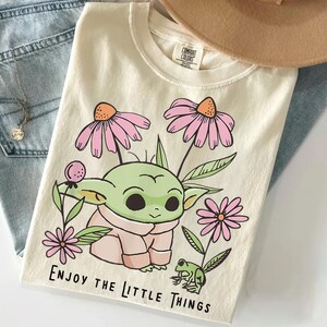 May include: A cream-colored t-shirt featuring a graphic of a green creature with large eyes, surrounded by pink flowers and a small green frog. The text "ENJOY THE LITTLE THINGS" is printed in black at the bottom. The shirt has a crew neckline.