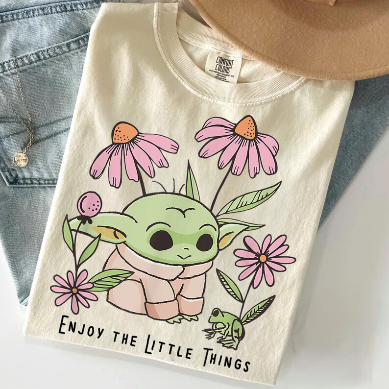 May include: A cream-colored t-shirt featuring a graphic of a green creature with large eyes, surrounded by pink flowers and a small green frog. The text "ENJOY THE LITTLE THINGS" is printed in black at the bottom. The shirt has a crew neckline.