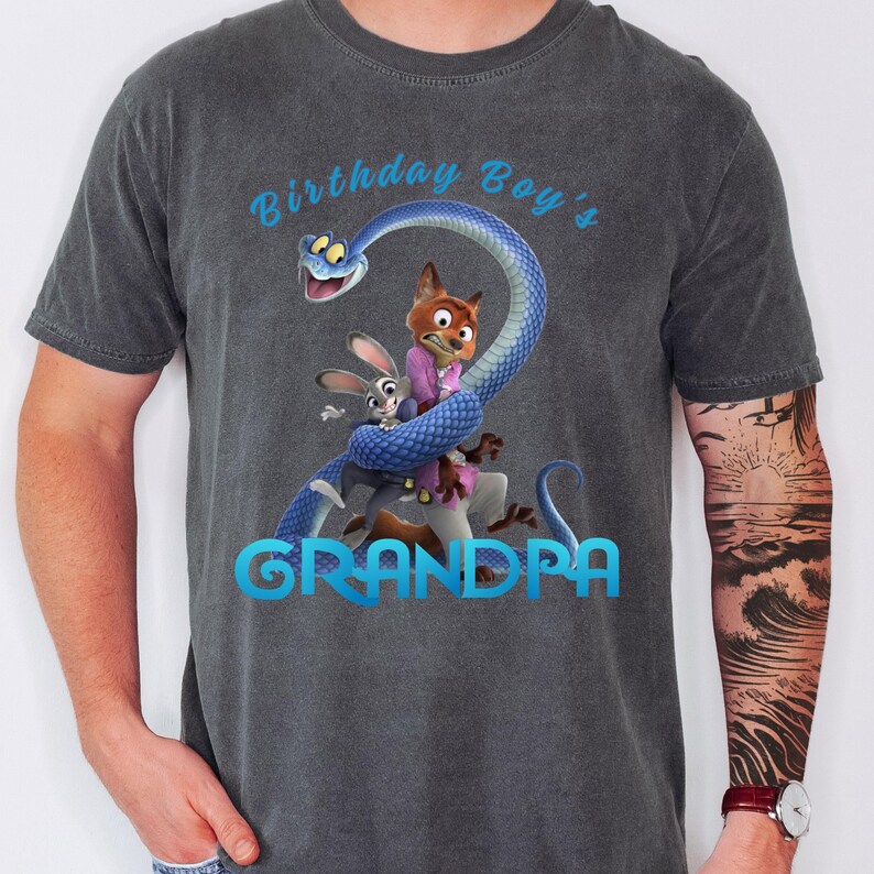 May include: A charcoal gray short-sleeve t-shirt featuring a graphic print of a blue snake coiled around a fox and a rabbit. The text "Birthday Boy's" appears in light blue above the characters, and the word "GRANDPA" is printed in light blue below.