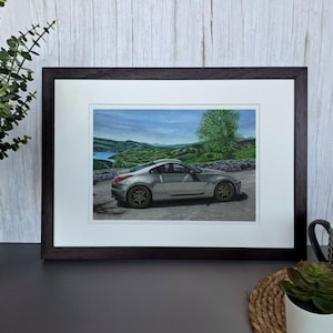 May include: A framed print featuring a silver sports car parked on a paved road with a scenic landscape of green hills, a body of water, and a blue sky in the background. The artwork is enclosed in a dark brown wooden frame with a white mat border.