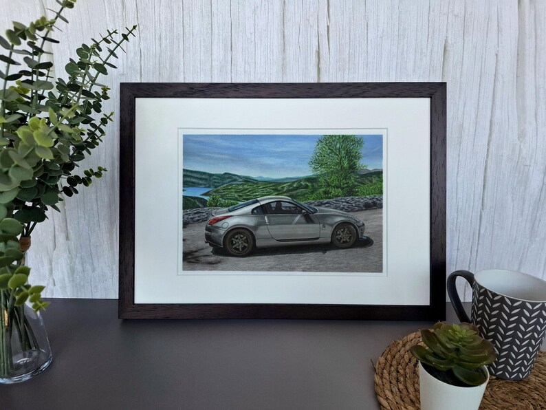 May include: A framed print featuring a silver sports car parked on a paved road with a scenic landscape of green hills, a body of water, and a blue sky in the background. The artwork is enclosed in a dark brown wooden frame with a white mat border.