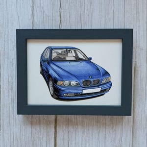 May include: A framed illustration of a blue BMW sedan shown from a front three-quarter angle. The car features a signature kidney grille, clear headlights, and a blank white license plate. The artwork is enclosed in a simple black rectangular frame.