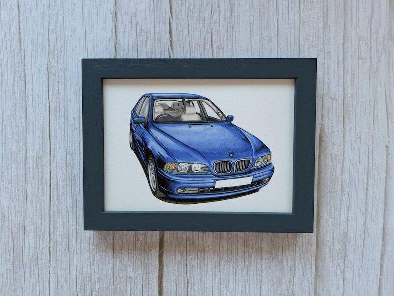 May include: A framed illustration of a blue BMW sedan shown from a front three-quarter angle. The car features a signature kidney grille, clear headlights, and a blank white license plate. The artwork is enclosed in a simple black rectangular frame.