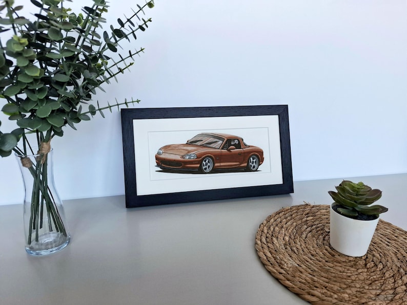 May include: A framed horizontal print featuring a detailed illustration of a metallic copper-colored convertible sports car. The artwork is encased in a simple black rectangular frame with a white mat border, showcasing the vehicle's sleek profile.