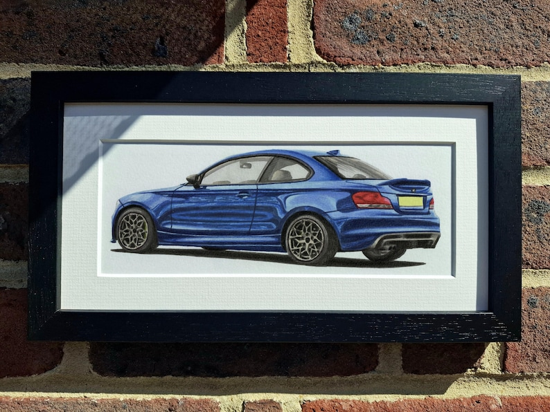 May include: A framed illustration of a blue coupe car shown from a rear three-quarter angle. The vehicle features dark multi-spoke wheels, a rear spoiler, and a yellow license plate. The artwork is enclosed in a simple black rectangular frame.