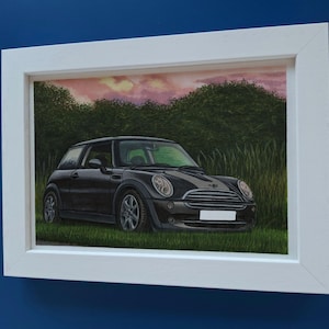 May include: A framed painting of a black Mini Cooper parked in a grassy field. The car is positioned in profile against a backdrop of green trees and a pinkish sunset sky. The artwork is enclosed in a simple white rectangular frame.