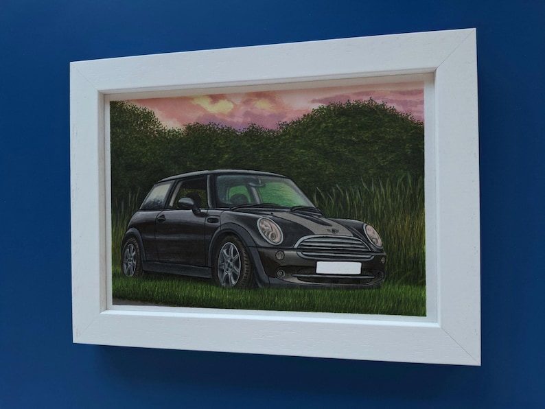 May include: A framed painting of a black Mini Cooper parked in a grassy field. The car is positioned in profile against a backdrop of green trees and a pinkish sunset sky. The artwork is enclosed in a simple white rectangular frame.