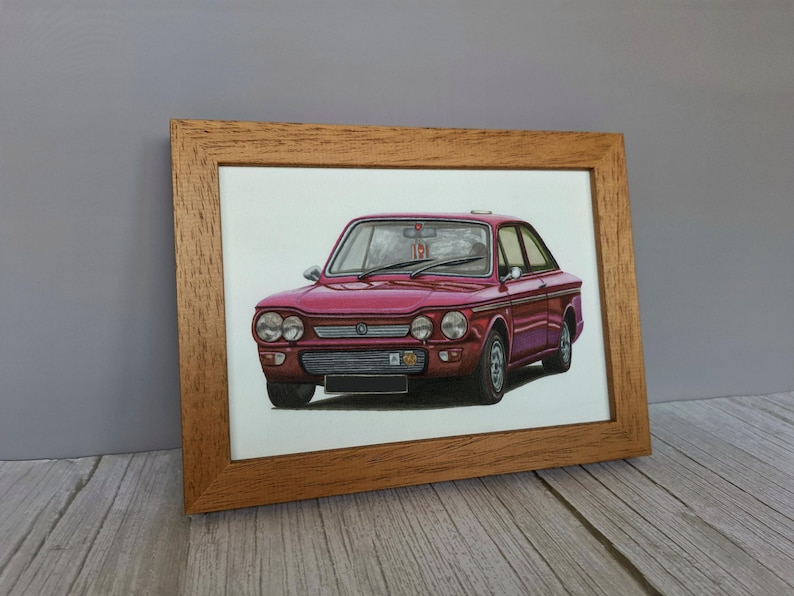 May include: A framed illustration of a vintage maroon two-door sedan. The car features a horizontal front grille, dual round headlights on each side, and a black license plate area. The artwork is encased in a light brown wooden frame.