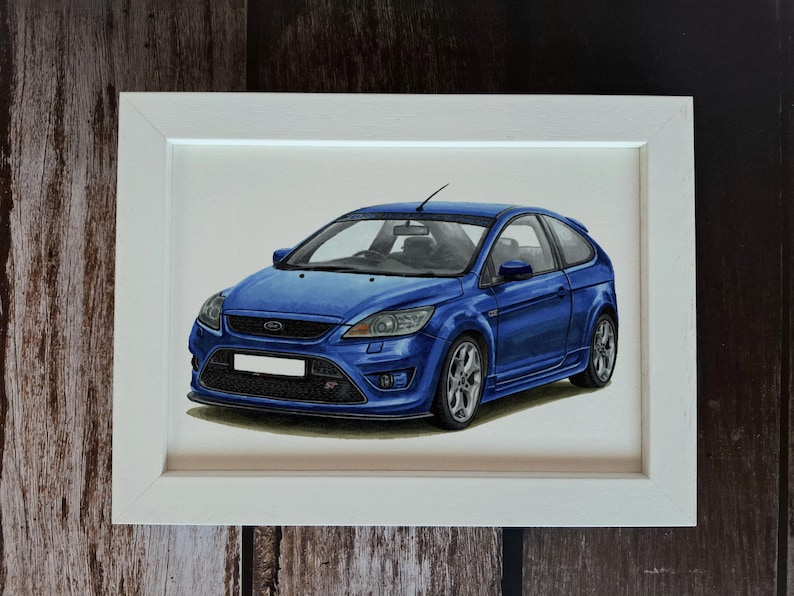 May include: A framed illustration of a blue Ford Focus ST hatchback. The car is depicted in a three-quarter front view, showcasing its signature grille, headlights, and silver alloy wheels. The artwork is enclosed in a simple white frame.