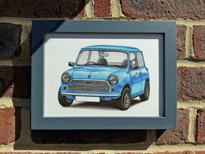 May include: A framed illustration of a light blue compact car featuring a horizontal slatted grille, round headlights, and a blank white license plate. The artwork is encased in a dark gray wooden frame with a clean, rectangular profile.