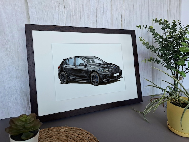 May include: A framed illustration of a black hatchback car shown from a side angle. The car features dark multi-spoke wheels, a sleek aerodynamic body, and a white license plate area. The artwork is presented in a dark brown wooden frame.