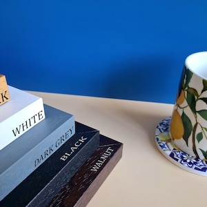 May include: A stack of five rectangular wooden frames labeled Oak, White, Dark Grey, Black, and Walnut, positioned next to a white ceramic mug featuring a yellow lemon and green leaf pattern resting on a blue and white patterned coaster.
