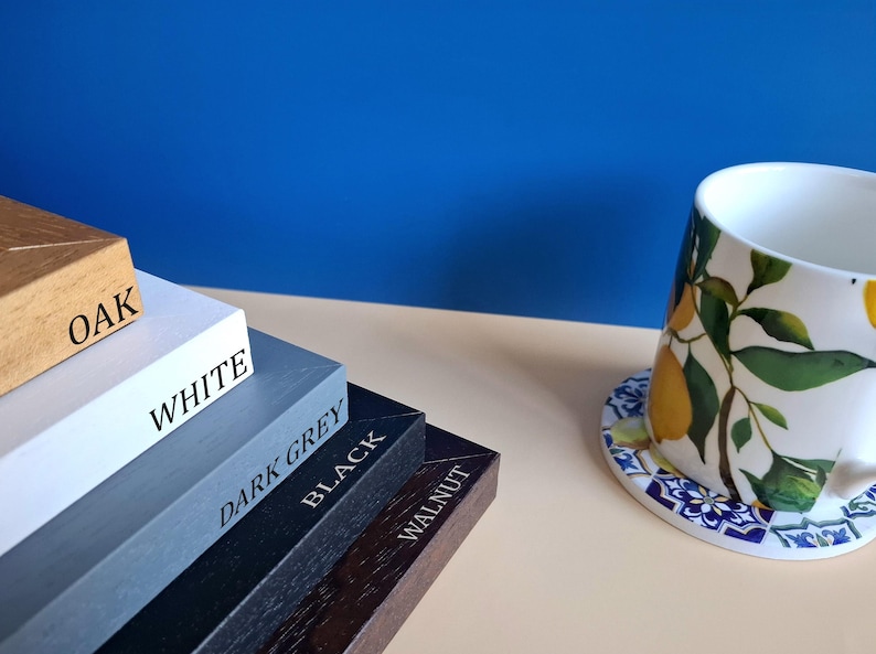 May include: A stack of five rectangular wooden frames labeled Oak, White, Dark Grey, Black, and Walnut, positioned next to a white ceramic mug featuring a yellow lemon and green leaf pattern resting on a blue and white patterned coaster.