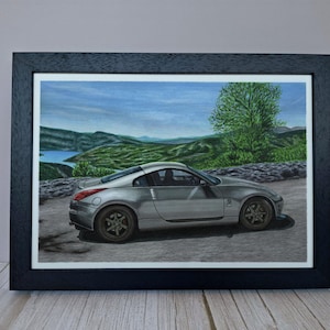 May include: A framed art print featuring a silver sports car parked on a paved road. The background displays a landscape with green hills, a body of water, and a large tree under a blue sky. The frame is black with a white mat border.