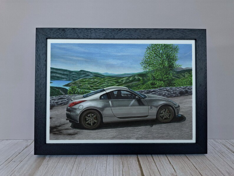 May include: A framed art print featuring a silver sports car parked on a paved road. The background displays a landscape with green hills, a body of water, and a large tree under a blue sky. The frame is black with a white mat border.