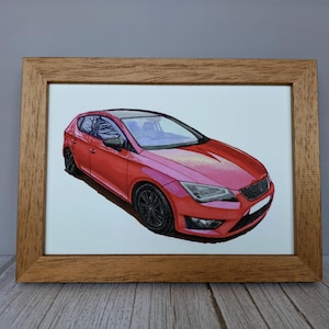 May include: A framed illustration of a red hatchback car with black wheels and a black roof. The car is depicted from a front three-quarter angle, showcasing its angular headlights, dark grille, and sleek body lines, all encased in a light brown wooden frame.