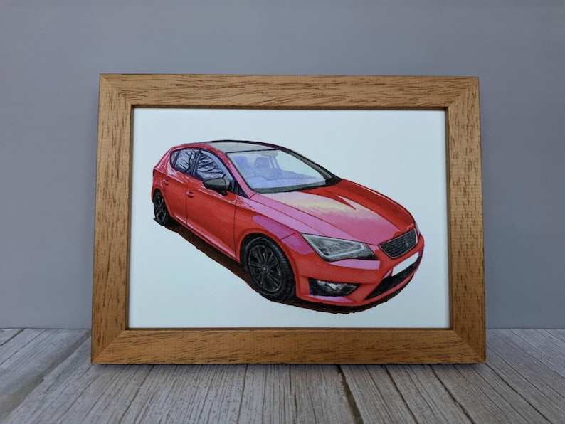 May include: A framed illustration of a red hatchback car with black wheels and a black roof. The car is depicted from a front three-quarter angle, showcasing its angular headlights, dark grille, and sleek body lines, all encased in a light brown wooden frame.