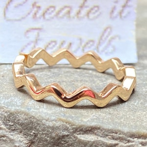 Può includere: Anello dorato con un design a zig-zag. L'anello è su una superficie testurizzata di colore marrone chiaro. Il testo "Create it Jewels" è visibile su una carta bianca dietro l'anello.