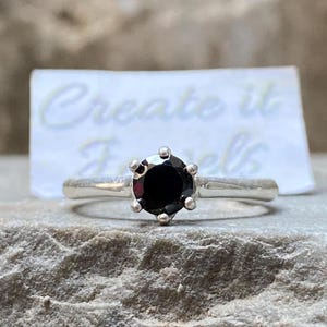 May include: A silver ring with a black gemstone set in a prong setting. The ring is on a gray rock surface. The text "Create it Jewels" is visible on a white card behind the ring.