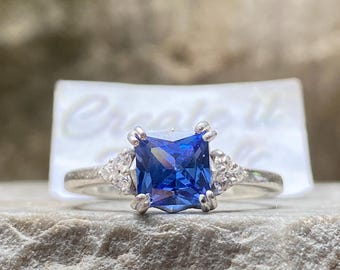 Op de afbeelding: Een zilveren ring met een grote, vierkante, diepblauwe edelsteen als middelpunt. Twee kleinere, heldere edelstenen flankeren de hoofdsteen. De ring is geplaatst tegen een natuurstenen achtergrond. De tekst "Create it" is zichtbaar.