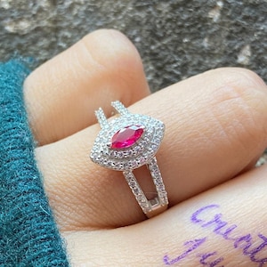 May include: A silver ring with a red marquise-shaped gemstone surrounded by smaller clear gemstones. The ring has a double band design.
