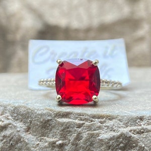 May include: A red gemstone ring with a square-shaped stone set in a gold band. The ring is on a grey rock surface.