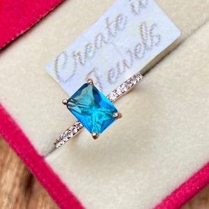 May include: A ring with a large, rectangular, blue gemstone surrounded by small, clear gemstones. The ring is in a pink velvet box with the text "Create it Jewells" on a white label.