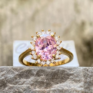 May include: A gold ring featuring a large, oval-cut pink gemstone surrounded by smaller, clear stones. The ring's band is a simple, polished gold. The setting of the ring is a halo design, with the smaller stones encircling the central pink stone.