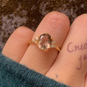 May include: A gold ring with a large oval-shaped, light brown gemstone as the centerpiece. The ring is flanked by smaller, clear gemstones on each side. The words "Create it jewels" are written on the finger.