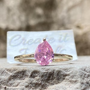 May include: A gold ring with a pear-shaped pink gemstone. The ring is on a gray stone surface.