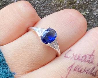 Op de afbeelding: Een zilveren ring met een grote, ovaal geslepen, diepblauwe edelsteen als middelpunt. De ring heeft aan weerszijden van de hoofdsteen een driehoekig ontwerp, elk met een kleinere, heldere steen. De woorden "Create it Jewels" staan op de vinger.
