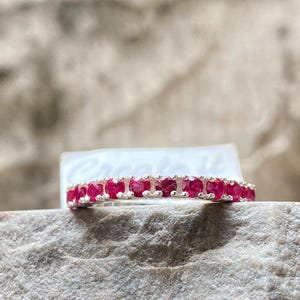 May include: A silver ring with a row of vibrant red gemstones. The ring is set against a textured, light gray stone background, highlighting the jewelry's sparkle and color. The gemstones are evenly spaced, creating a continuous band of red.