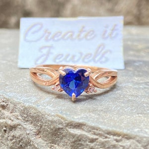 May include: A rose gold ring with a blue heart-shaped gemstone and smaller clear gemstones on the band. The ring is on a textured brown surface. The text "Create it Jewels" is visible on a white card behind the ring.