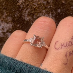 May include: A silver ring with a square, light brown gemstone and two smaller triangular stones on either side. The ring is on a finger with the words "Create it Jewel" written on it in purple ink. The background is a textured, dark surface.