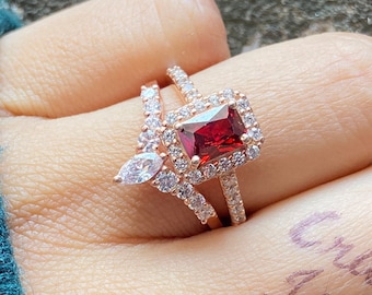 Op de afbeelding: Een ring van roségoud met een grote, rechthoekige, rode edelsteen omringd door kleinere, heldere edelstenen. De ring wordt gedragen aan de vinger van een hand.