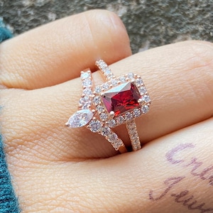 May include: A rose gold ring set with a large, rectangular, red gemstone surrounded by smaller clear gemstones. The ring is worn on the finger of a hand.