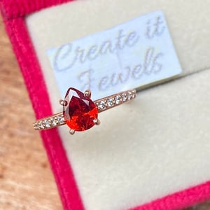 Peut inclure: Une bague en forme de poire avec une pierre rouge et une bande sertie de diamants. La bague est dans un écrin en velours rose avec l'inscription "Create it Jewels" sur une étiquette blanche.