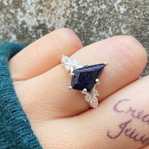 Könnte beinhalten: Ein silberner Ring mit einem dunkelblauen Edelstein, der in einer geometrischen Form gefasst ist. Der Ring ist mit kleinen weißen Steinen verziert und hat ein zartes, blattartiges Design.