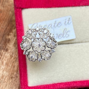 Puede incluir: Un anillo de plata con un diseño floral con múltiples piedras pequeñas transparentes. El anillo está en una caja de anillo de terciopelo rojo con el texto "Create it Jewels" en una etiqueta blanca.