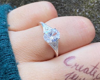 Peut inclure: Bague en argent ornée d'une grosse pierre précieuse ovale et transparente. La bague présente un design délicat avec de petites pierres d'accent. Le mot "Create it jewel" est écrit sur le doigt.