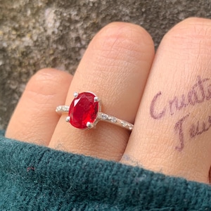 May include: A silver ring with a large oval ruby gemstone. The ring is set with smaller accent stones along the band. The words "Create it Jewel" are written on the fingers.