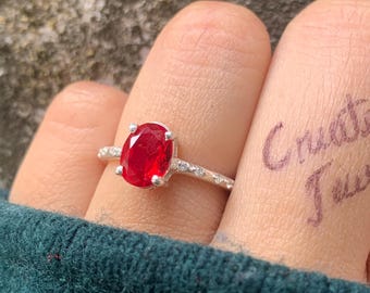 May include: A silver ring with a large oval ruby gemstone. The ring is set with smaller accent stones along the band. The words "Create it Jewel" are written on the fingers.