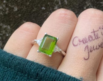 May include: A silver ring featuring a rectangular, light green gemstone as the centerpiece, flanked by two smaller, triangular clear stones. The ring is set against a textured background. The words "Create it Jewel" are written on a finger.