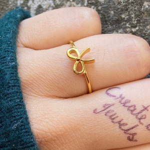 May include: A gold-colored ring with a bow design. The ring is worn on the index finger of a hand.